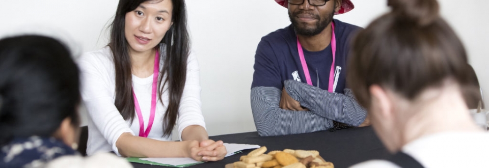 Sandy and Nolan are sat at a table, both listening in conversation to someone off-camera. Sandy is wearing a white top. Nolan is wearing a dark t-shirt with a grey shirt underneath, and a bucket hat.