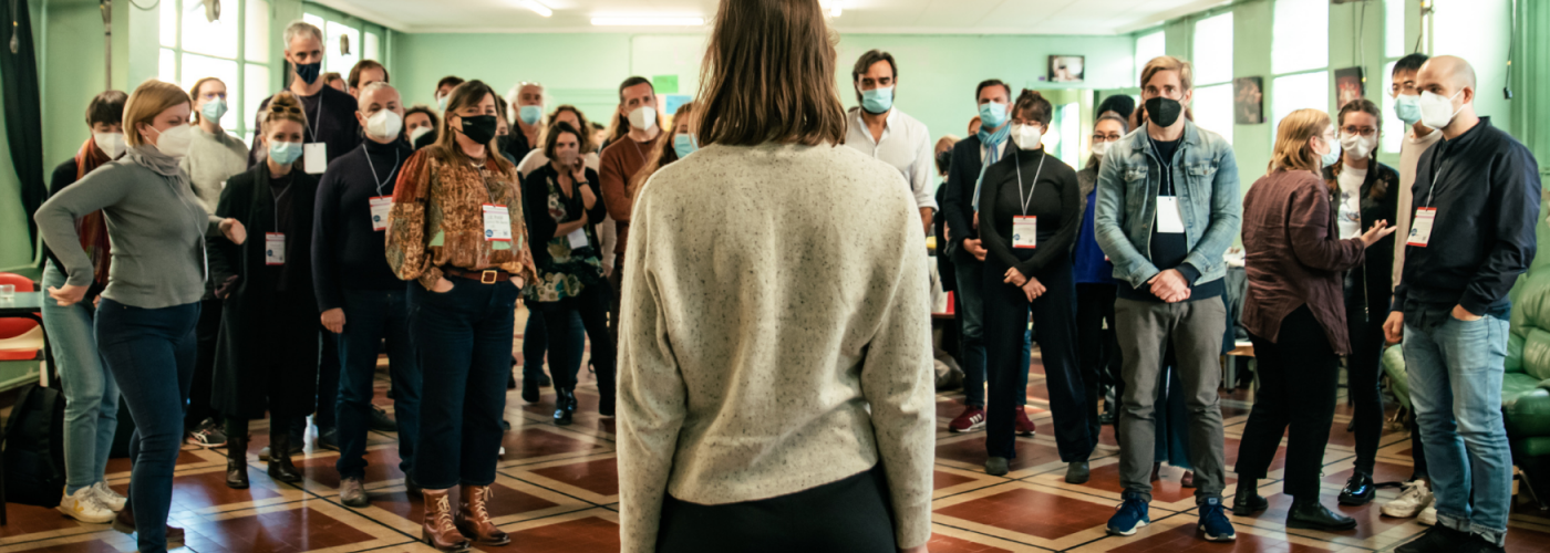 The banner displays a woman with her back turned to the camera, facing a crowd of people with masks in a room of the IETM Lyon Plenary Meeting 2021