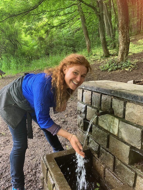 woman with red hair in a blue shirt bending with hand outstretched into spring water flowing from a man made well