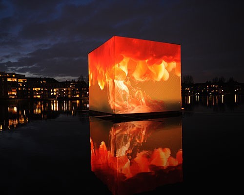 ThThe CO2 Cube: Visualizing a Tonne of Change a brilliant red and orange cube lit by projections of fire, floating in water on a dark night, Copenhagen 2009dark night