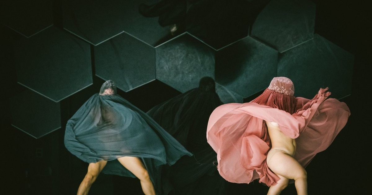 Corpo Clandestino The imcage shows two bodies, half covered in different transparent fabrics, that soflty fly around them. Behind the two bodies, a third one covered in black fabric is visible. Their heads and faces are covered. They dance in front of a grey, green background.