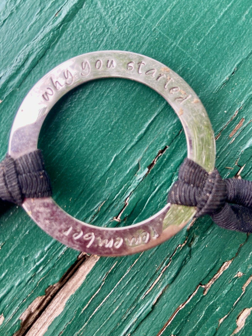 round metal ring with two black cords sat on a green peeling   painted wooden surface - 'remember why you started' etched into the metal