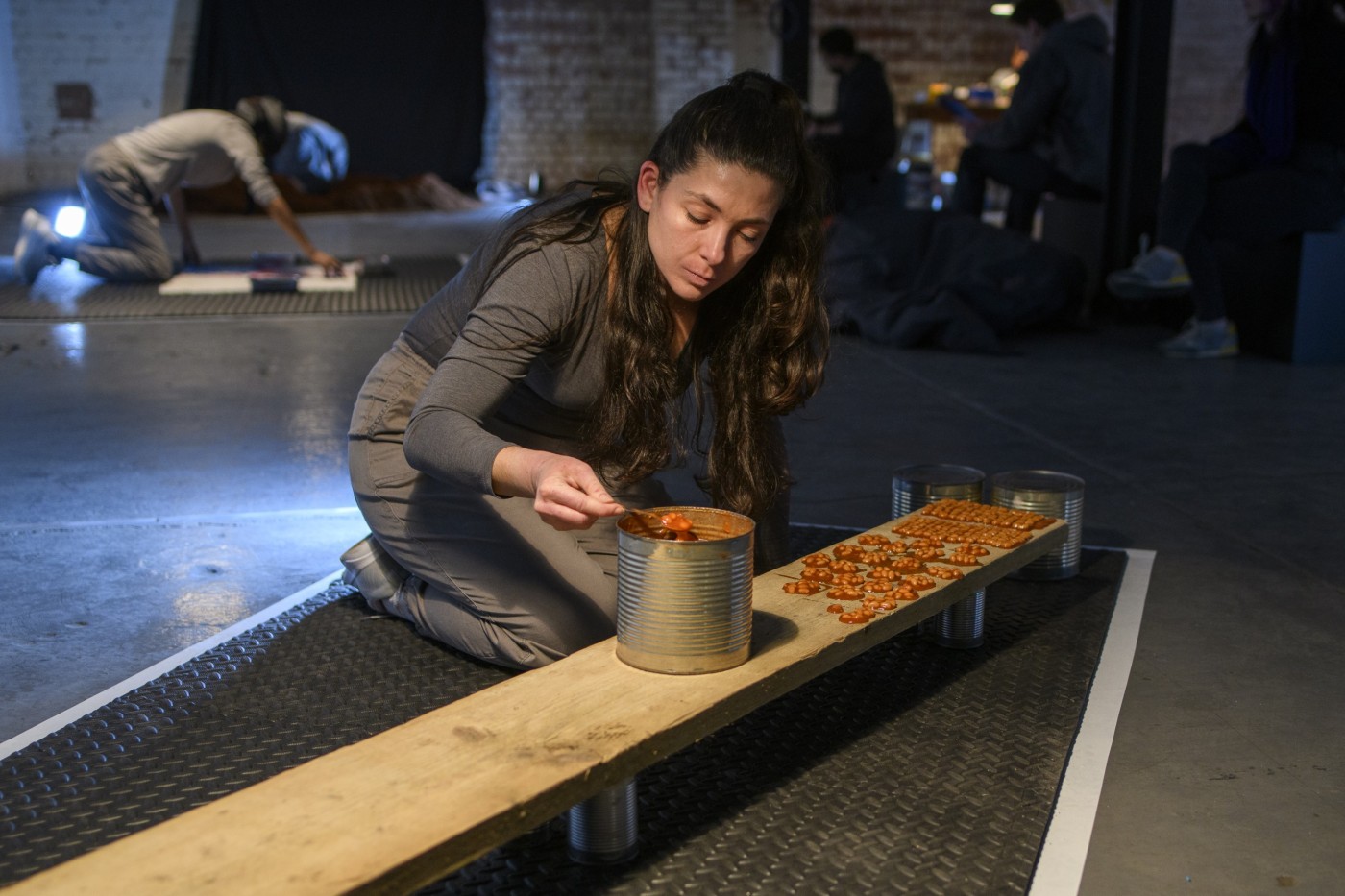 A woman is kneeling by a plank of wood. She is taking baked beans from a big tin with a spoon. There are more baked beans arranged in patters on the plank in from of her. 