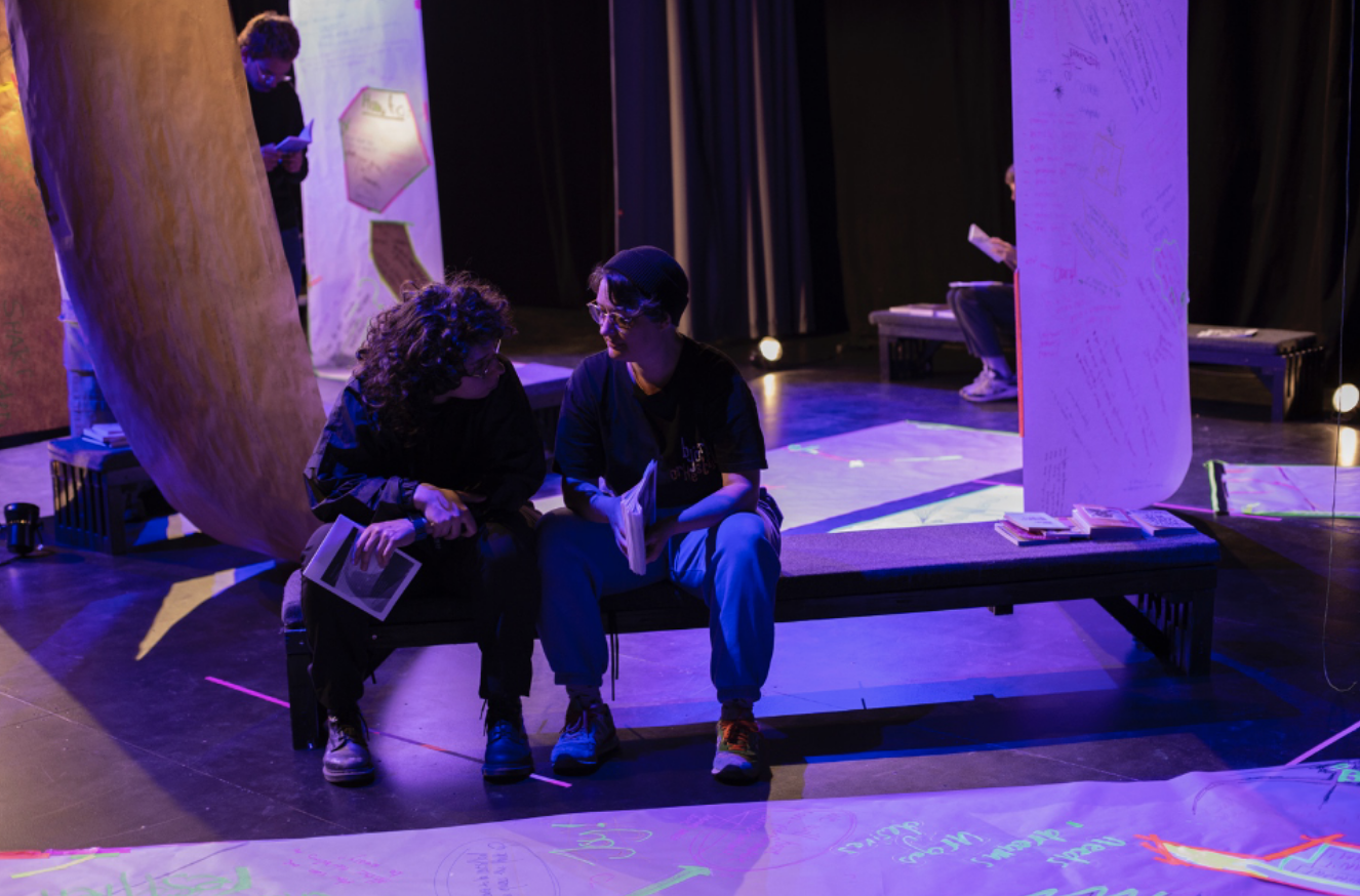 Zee + Joni are sitting on a bench inside a blackbox. Paper lines are hanging in the room. The light is purple, blue.