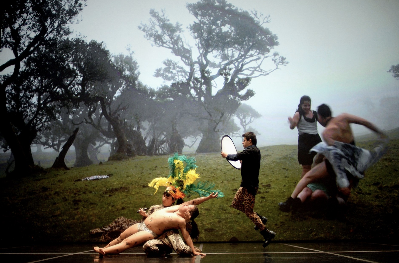 three dancers are on the stage in front of a screen, that shows a hill covered in grass, trees in the back. There also 4 bodies are dancing. 