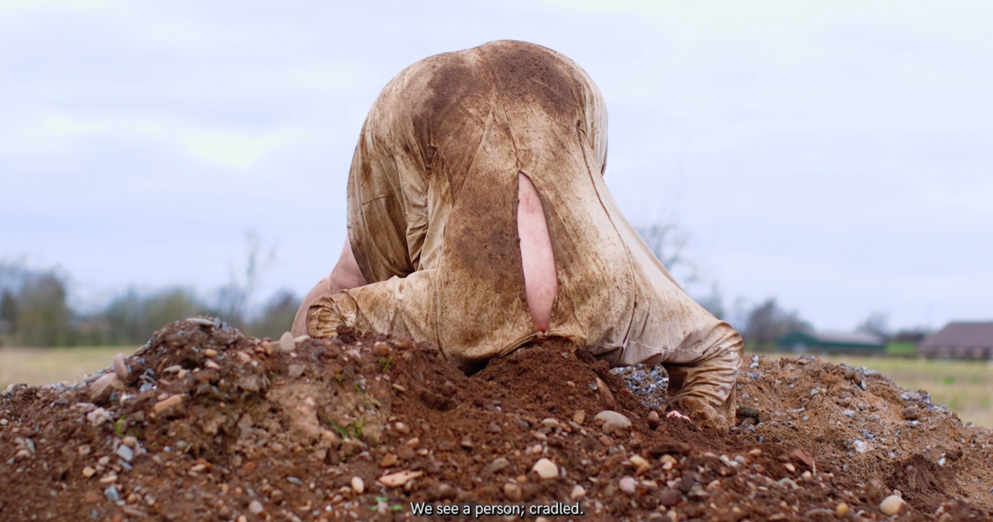 A white person is upside down, their head buried in a large pile of soil. Their clothing is made of natural materials but also muddy. Beyond them the landscape is great with an overcast blue sky.