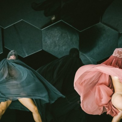 The image shows two bodies covered in transparent fabric, one dark green, the other in light pink, that float around the dancers bodies. In the background, a third body covered in transparent black fabric can be sensed. Their heads and faces are covered. They dance in front of a dark green background. 