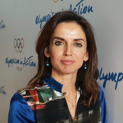 Head and shoulders image of a white woman with long brown hair wearing a colourful shirt standing in front of a whiteboard with writing on it