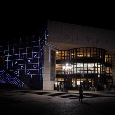 Night view of Divadlo Andreja Bagara in Nitra with a projection of a grid and tic-tac-toe symbols on its facade, part of the Divadelná Nitra festival programme, visitors gathered on the square in front of the building.