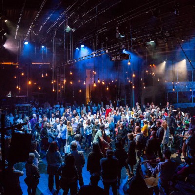 A photography of an audience standing on a stage lit by hanging light bulbs