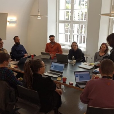 The photo represents a group of 10 people, members of On the Move discussing around a table. 